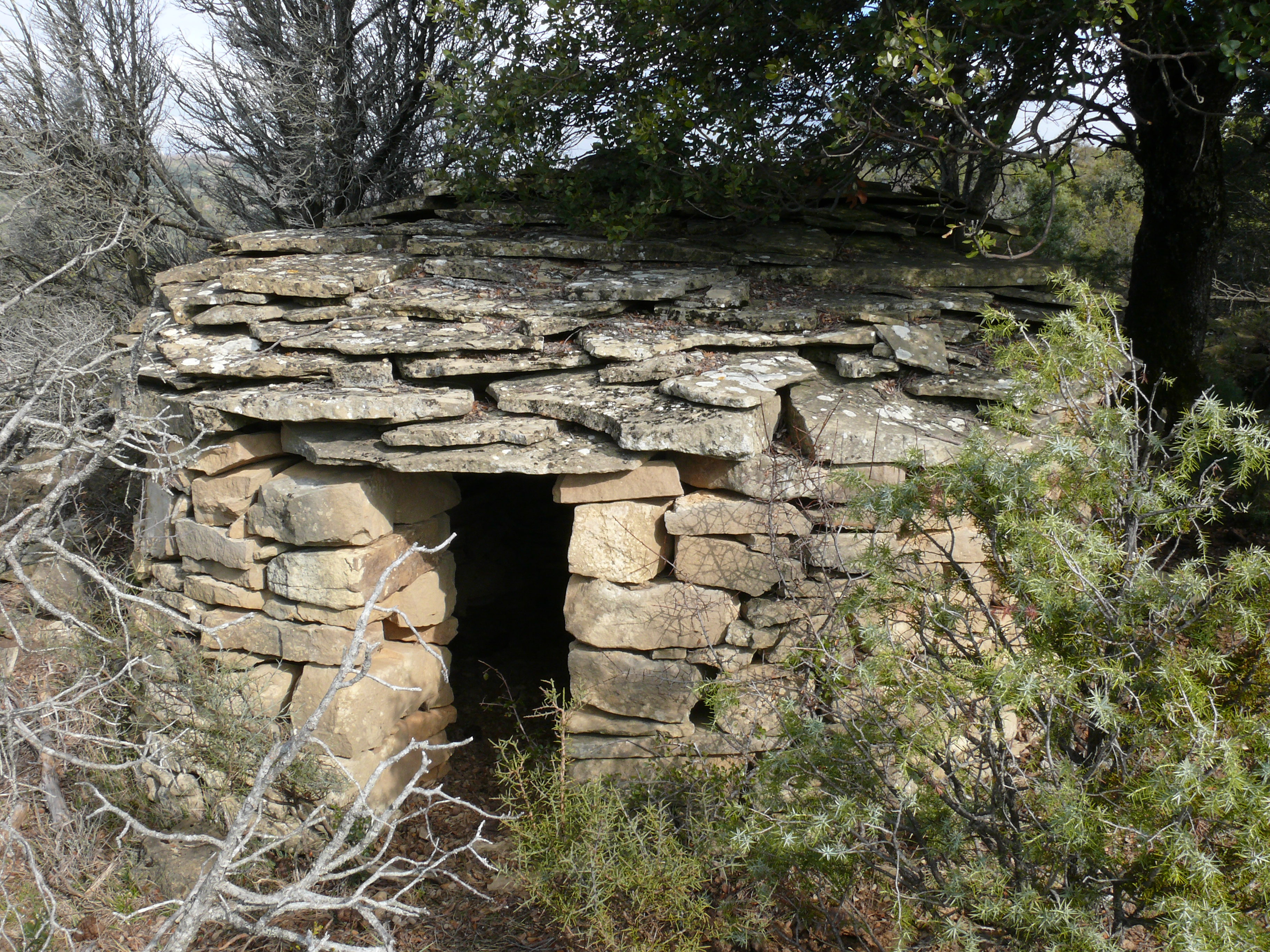 Vista de la cabaña, de planta circular, con tejado de losa de piedra, a una sola agua.